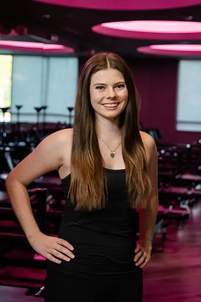 Hailey, Lagree instructor, smiling with hands on hips in a pink-lit studio.