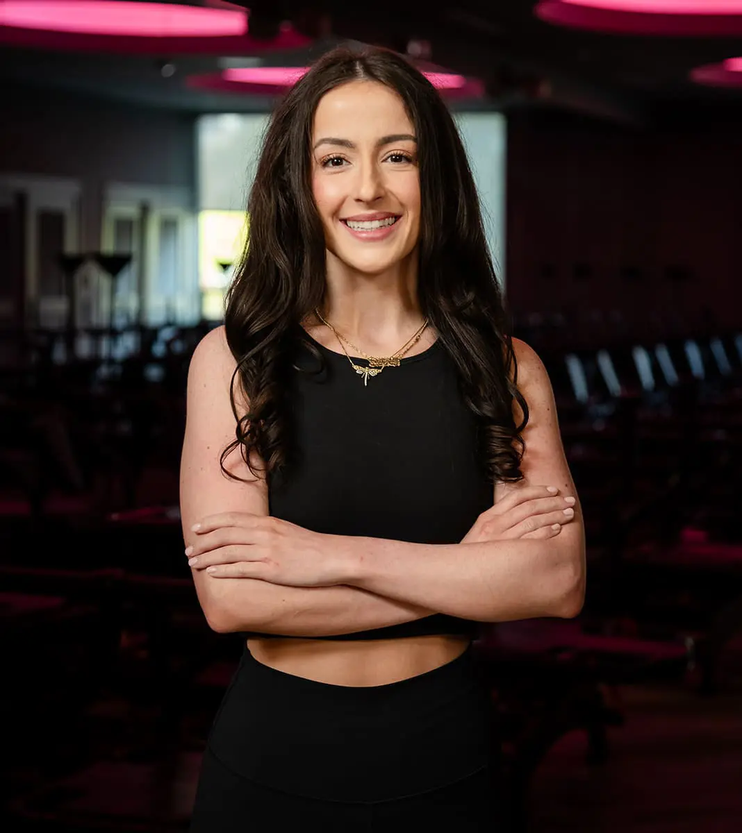 sydneigh-fitness-instructor-portrait-with-arms-crossed-in-red-lit-studio-tight-dallas. Sydneigh, fitness instructor, standing with arms crossed and smiling in a dimly lit Lagree studio.