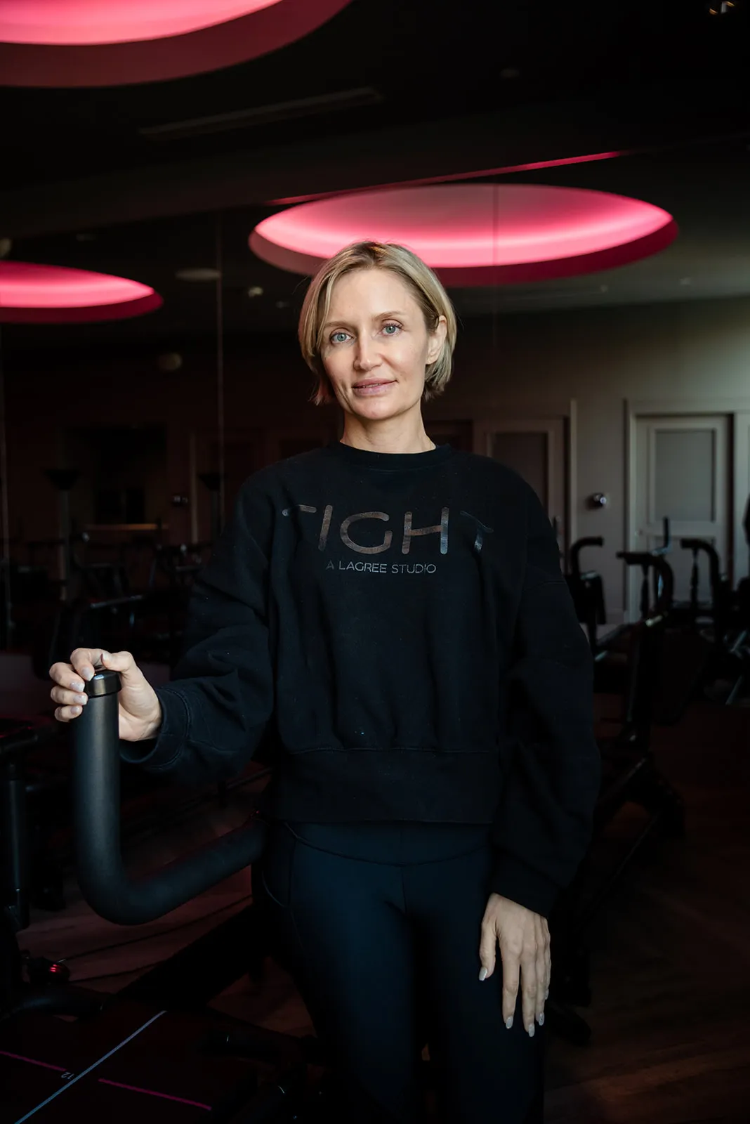 trainer-portrait-beside-megaformer-tight-dallas. Trainer in a branded TIGHT sweatshirt stands beside a Megaformer in the Dallas Lagree studio